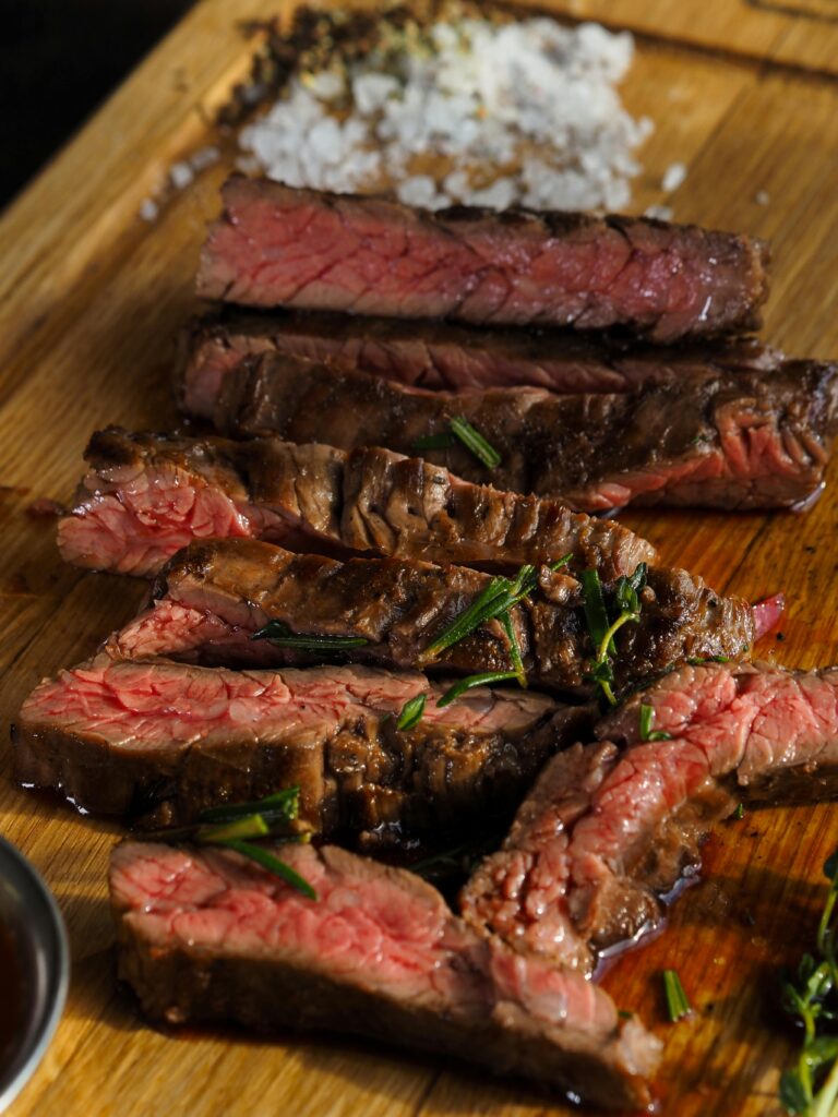 Close-up of sliced grilled steak with seasoning on a wooden board, highlighting texture and juiciness.